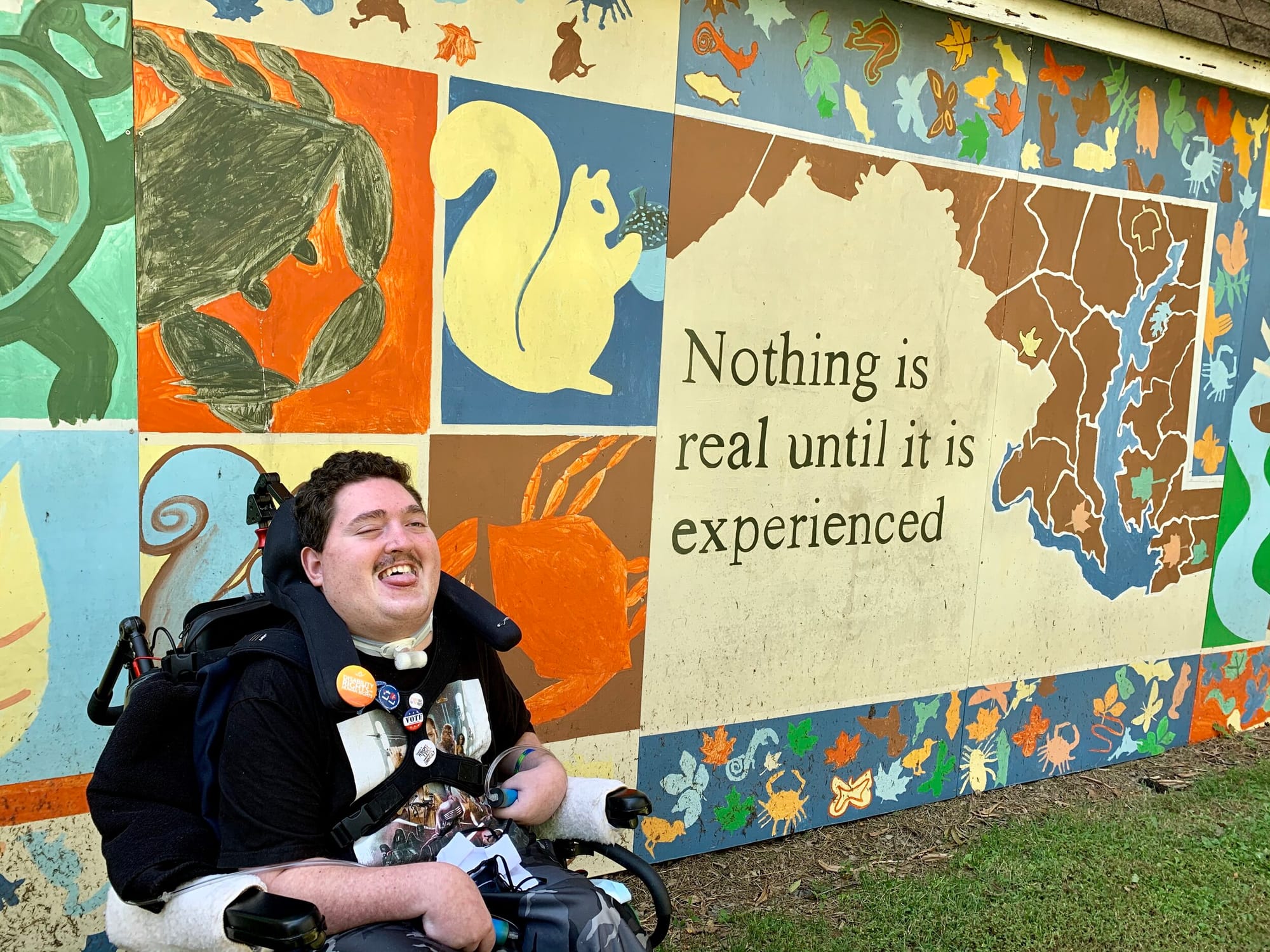 Rob at a recent state park outing. [A young man with light skin sits in his wheelchair in front of a colorful wall mural with a Maryland State Map, beneath which is written “Nothing is real until it is experienced.” There are also painted images of a turtle, a crab, and a squirrel visible.]
