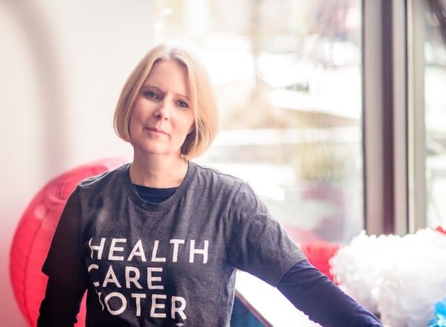 Angela Carpenter Gildner [image description: a white-appearing woman with blond hair poses in a gray “Health Care Voter” t-shirt with a red cushion in the background]
