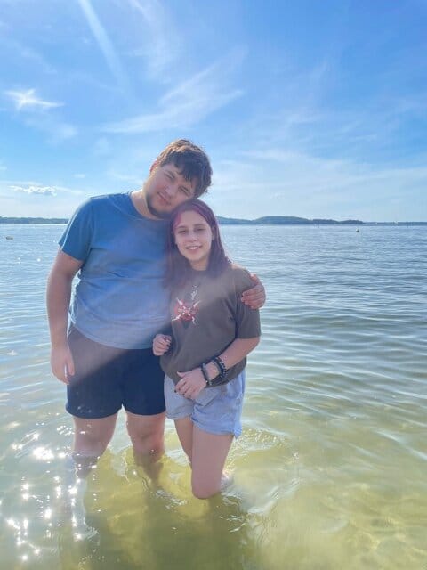 Graham and Margot [image description: a white-appearing brother and sister, each wearing t-shirt and shorts pose knee-deep in a lake with mountains in the distance on a sunny day]