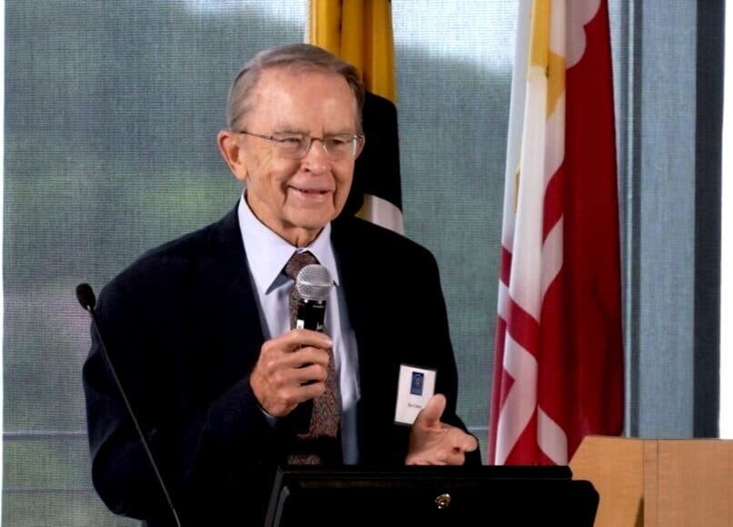 Ron Carlson [image description: A white-appearing man speaks at a lectern with a microphone]