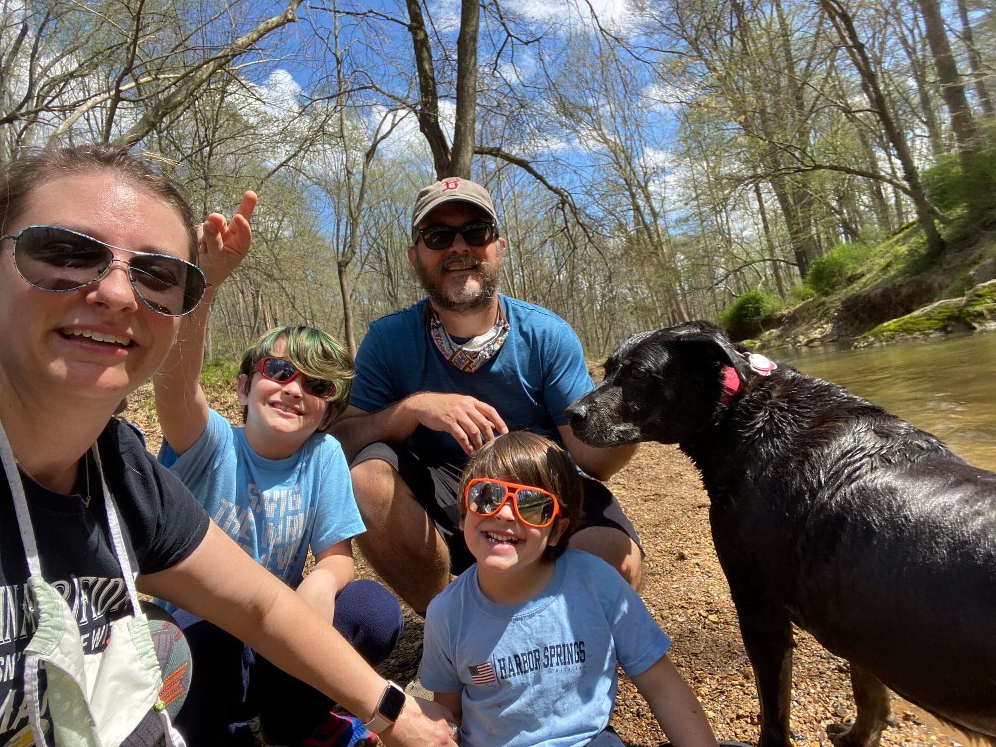 Peter Witzler and his family. [image description: A white-appearing father, mother and two young boys pose with their black dog in a wooded area near a stream. All are smiling.]