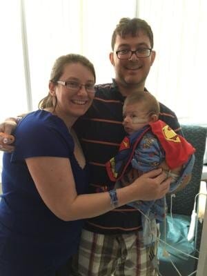 Peter and Lisa with Jackson just before his first surgery at three-and-a-half months. [image description: Two white-appearing parents hold their infant son, who wears a Superman cape.]