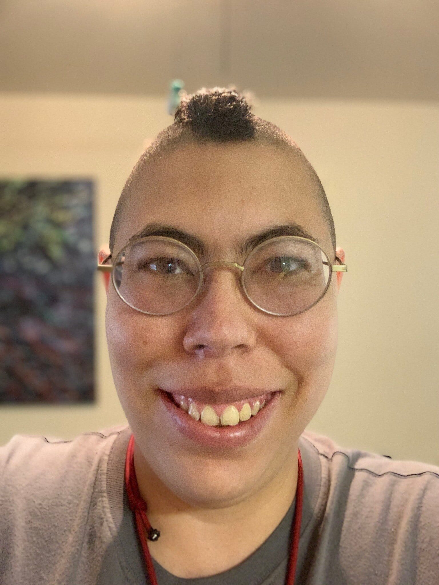 Angela Weddle [image description: A BIPOC person smiles into the camera. She wears round, metal-framed glasses and a grey shirt. Her hair is styled as a narrow strip of curly brown hair, with the rest of the head shaved on both sides.