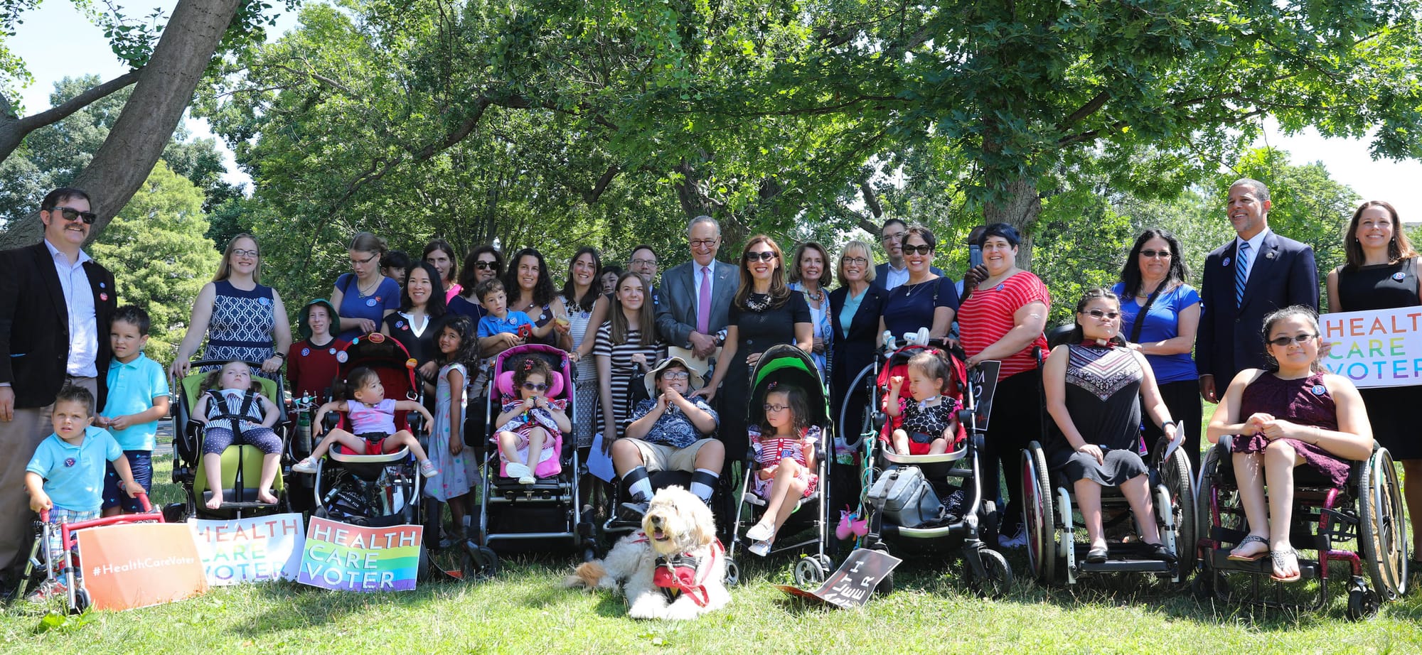 Members of Little Lobbyists pose with Senator Chuck Schumer on the Capitol Building grounds. [image description: A large group of adults and children, many using alternative mobility, pose in front of bright green summer tree foliage.]