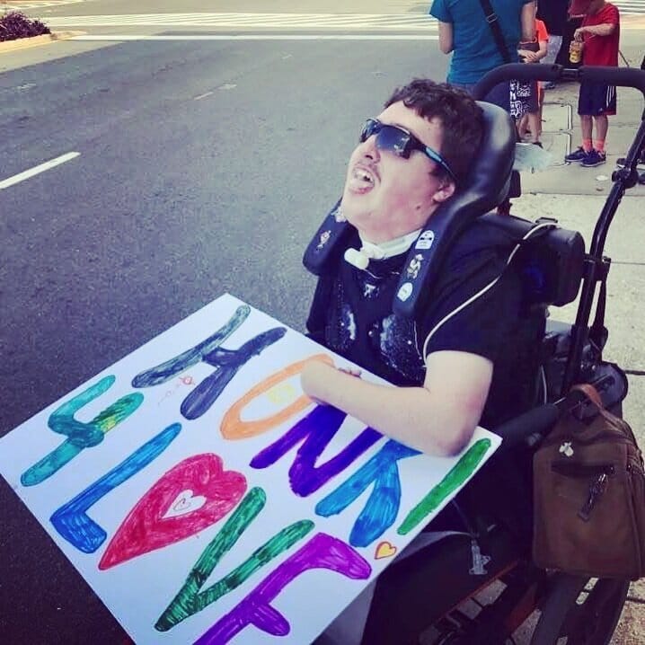 Rob Stone at a Bethesda protest against the violence in Charlottesville, Virginia. [image description: A young white man wearing sunglasses is seated in a wheelchair. He holds a multicolored, hand-painted sign, “HONK! 4 LOVE".]