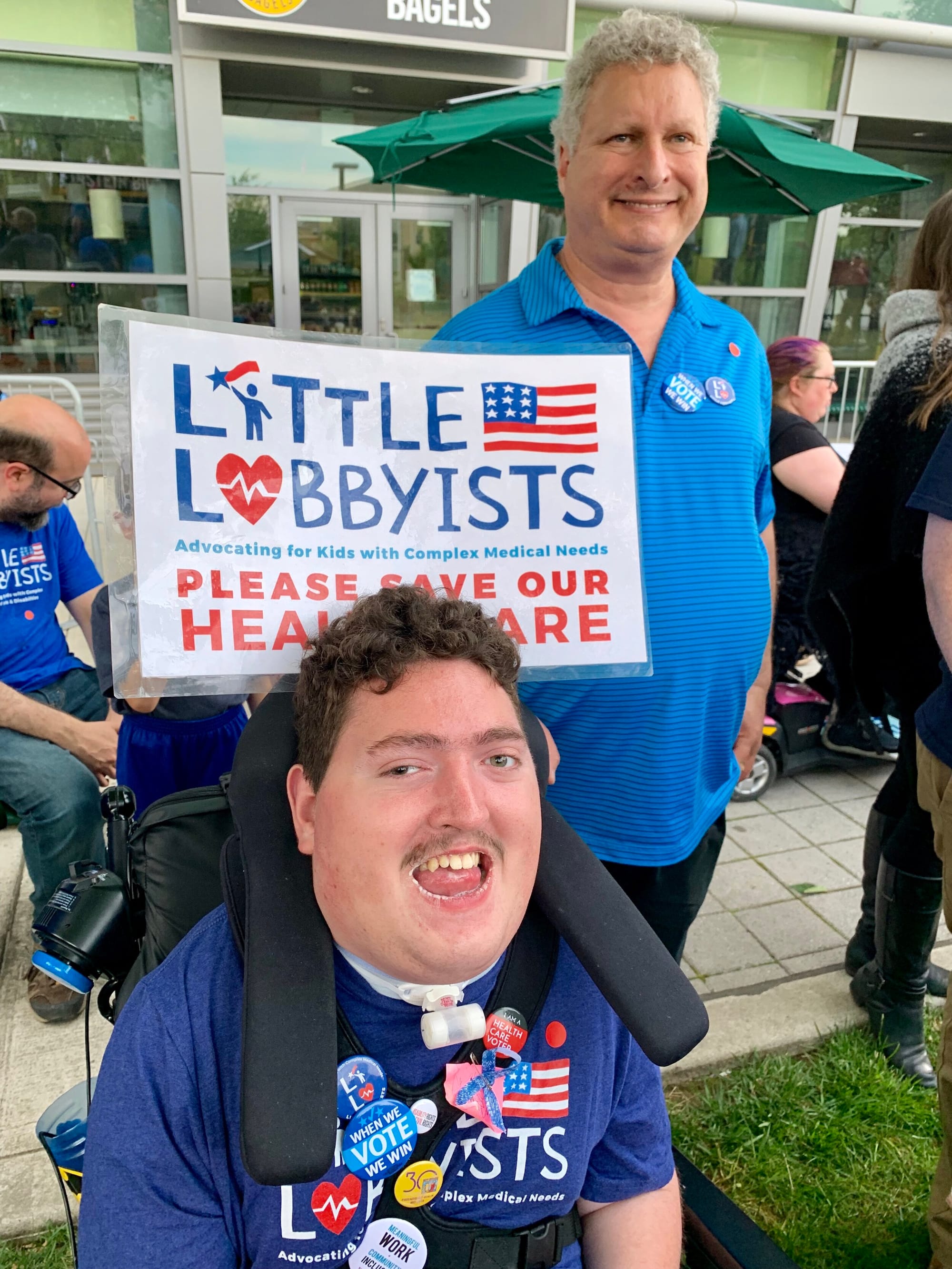 Rob Stone and his dad advocating with Little Lobbyists [image description: A young white man with short, curly dark hair, poses in his wheelchair, his supportive vest covered in political buttons. His head rest supports a sign that says, “Little Lob…