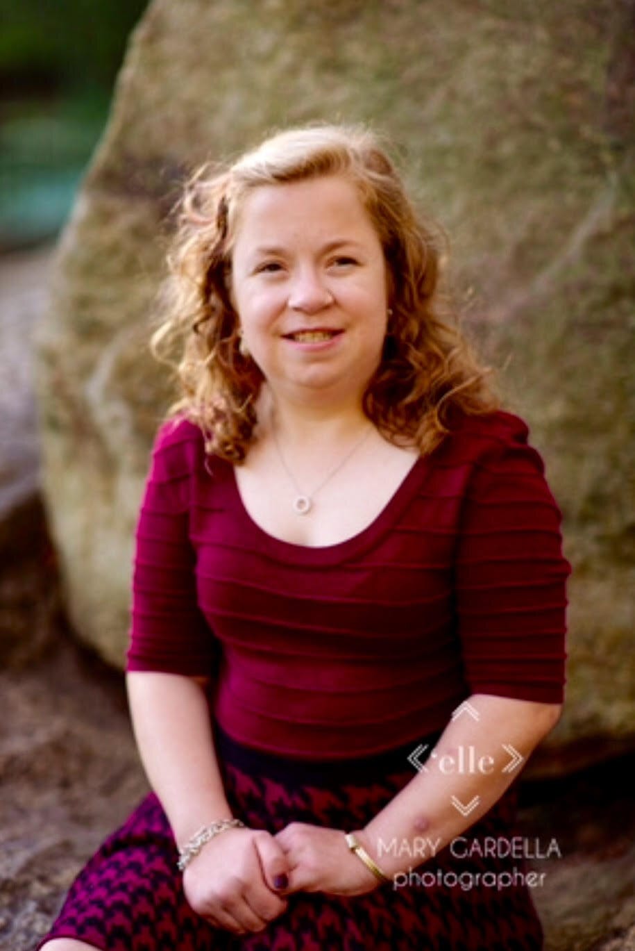 Sierra today. [image description: A young white woman with long curly blonde hair is seated outdoors. She wears a scoop-neck burgundy top and a burgundy and black checked skirt.]
