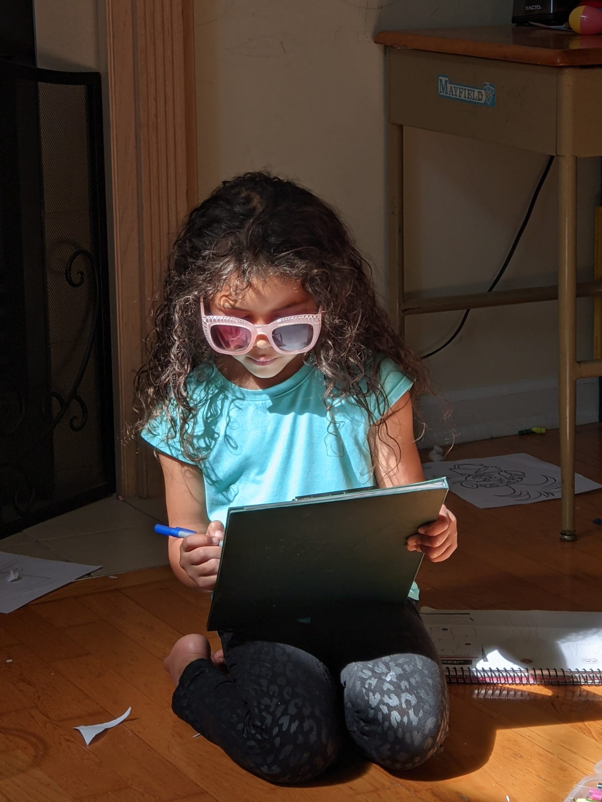 Maya’s daughter Ruby. [image description: A young Black girl with long, curly black hair wears pink sunglasses while she makes notes with a pen on a blue pad.]