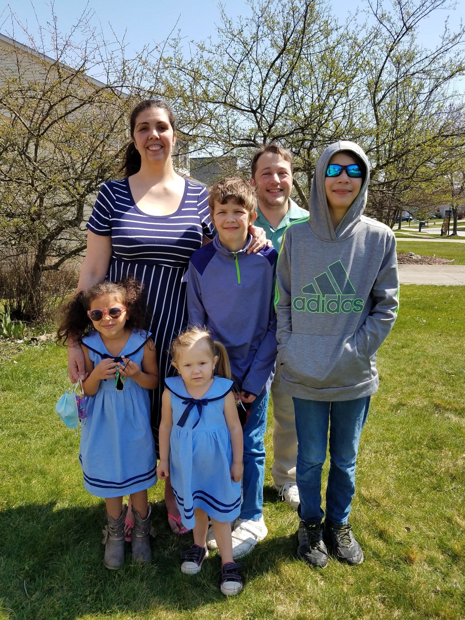 The Brown-Zimmerman Family. [image description: A family of two adults and four children poses on a green lawn with trees in the background that are just beginning to leaf out. The girls wear matching light blue sailor dresses, and the boys wear ath…