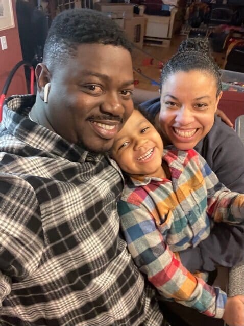 Amadou, Zana, and Amy. [image description: A Black family poses on a couch. The dad wears a black &amp; white plaid shirt, the son wears a colorful striped shirt, and the mom wears a gray shirt, her hair pulled back into a bun.]