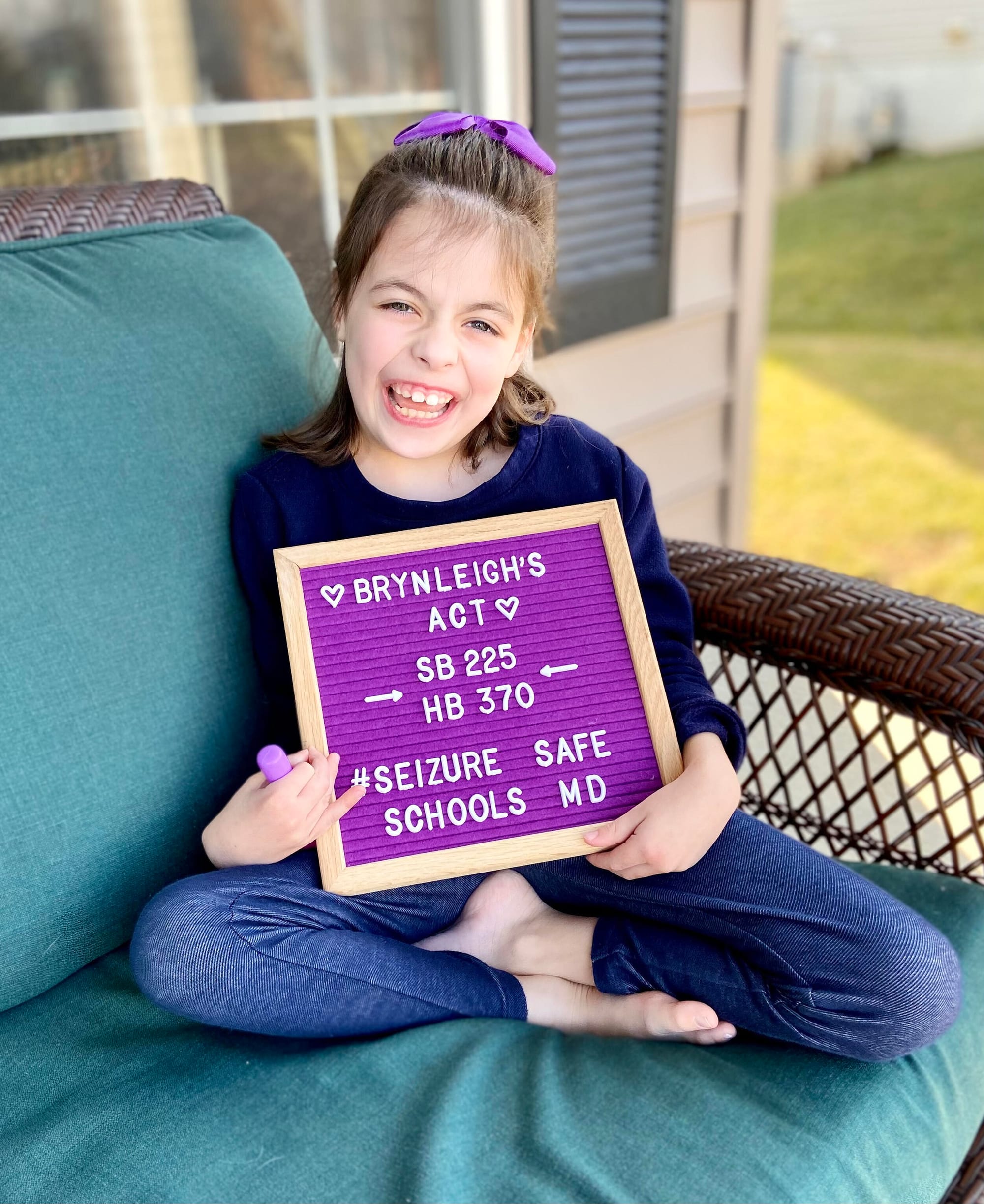 Brynleigh Shillinger [image description: A young girl with dark hair and a purple hair bow poses in a wicker outdoor chair with teal cushions. She holds a purple sign that reads “Brynleigh’s Act, SB225, HB370, #Seizure Safe Schools MD.”]