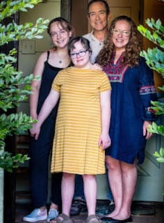 The Silverman-Stern Family: Annabelle, Sophie, Ray &amp; Amy [image description: A family poses at their front door, framed by green leaves of adjacent bushes.]