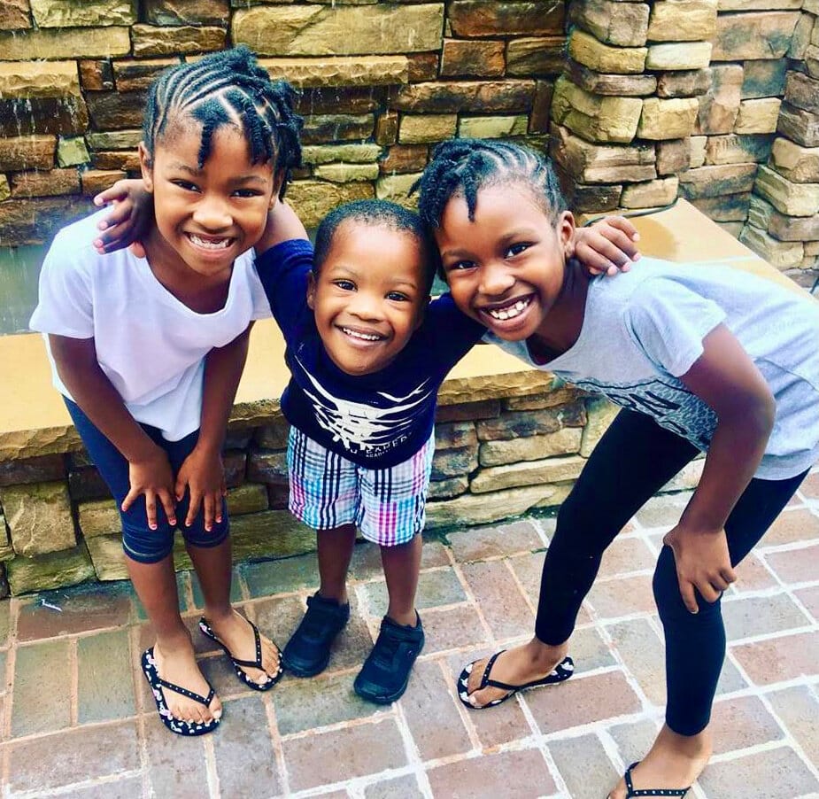 Anian &amp; his sisters. [image description: Standing in front of a fountain are two young Black girls with cornrow braids pose on either side of a young Black boy with Down Syndrome.]