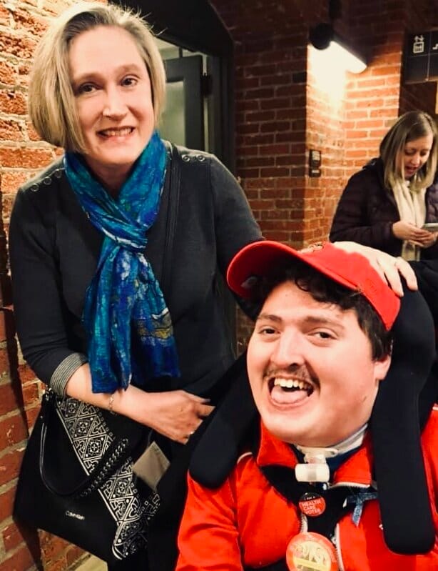 Jeneva &amp; her son Rob [image description: A white mom with short blonde hair wears a blue scarf &amp; grey dress. Her son is seated in a wheelchair with an orange shirt &amp; orange baseball cap.