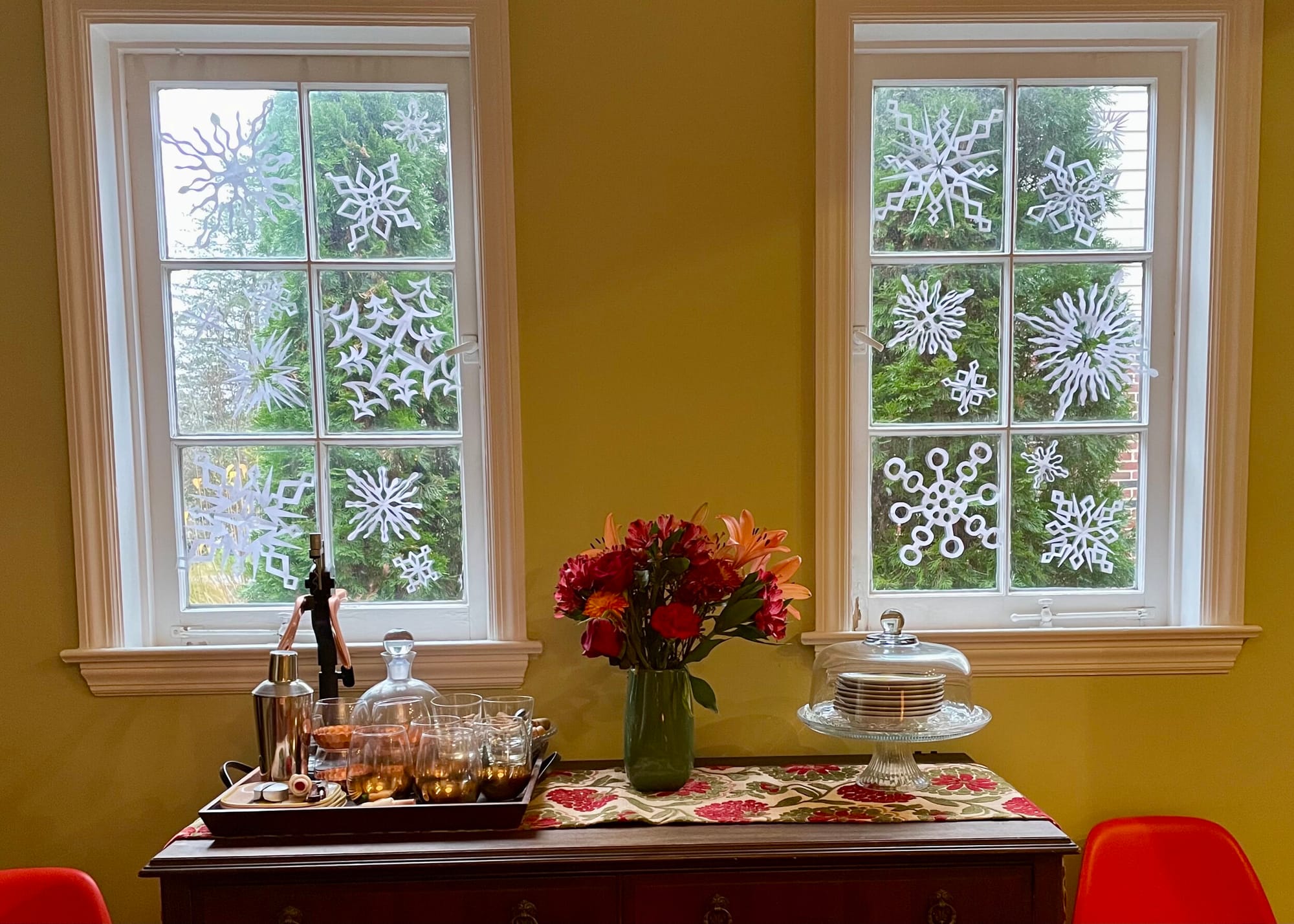 Laura’s snowflakes. [image description: Above a table set with glasses, flowers and dishes are two windows decorated with paper snowflakes.]