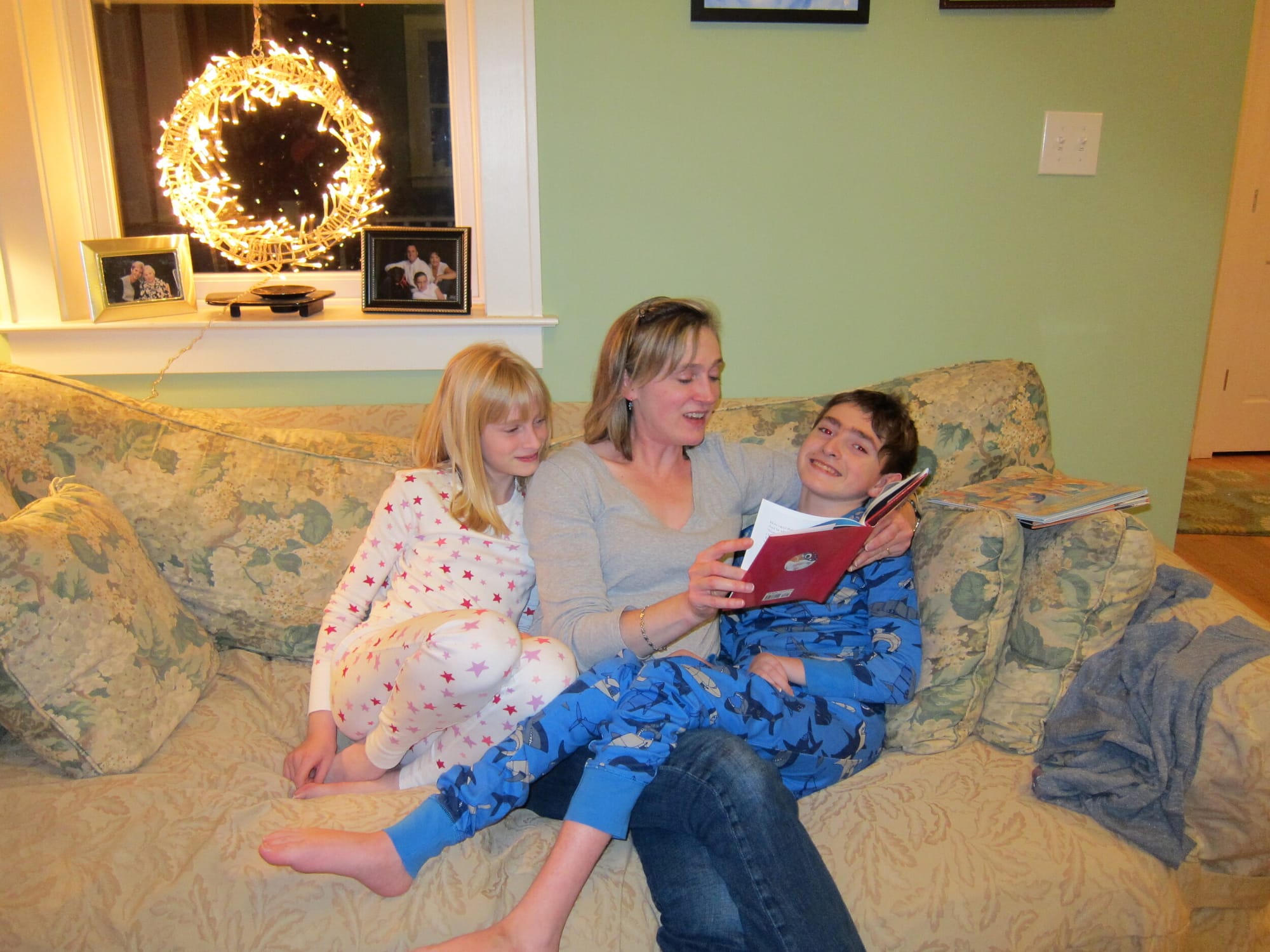Jeneva reads holiday stories to her children Castin (l) and Rob (r) in 2012. [image description: Seated on a couch, with a brightly lit wreath in a window above, a mom reads a book to two young children.]