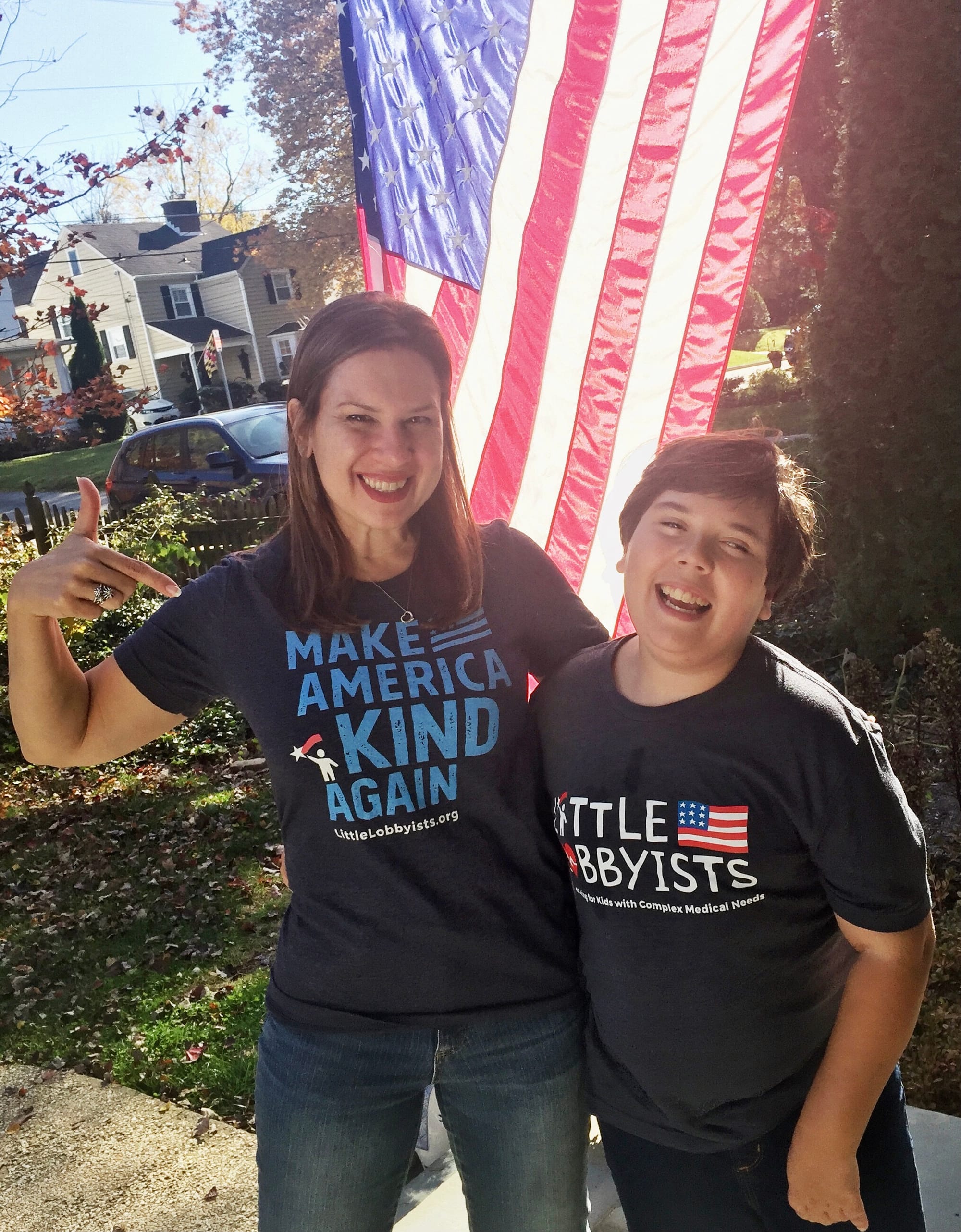 Laura and her son Simon. [image description: A mom and her son, both wearing nay blue Little Lobbyists t-shirts, stand in front of an American flag. The mom points proudly to her son.]