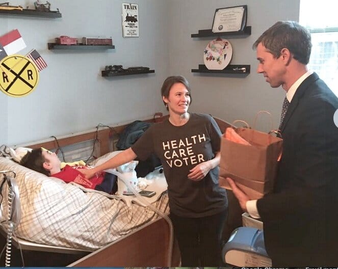 Laura Leeman stands next to Vic (who is in his bed) &amp; speaks with former congressman Beto O’Rourke. [image description: A woman with a “Health Care Voter” t-shirt faces a a man in a suit and tie. They are talking. Next to them, a young disabled …