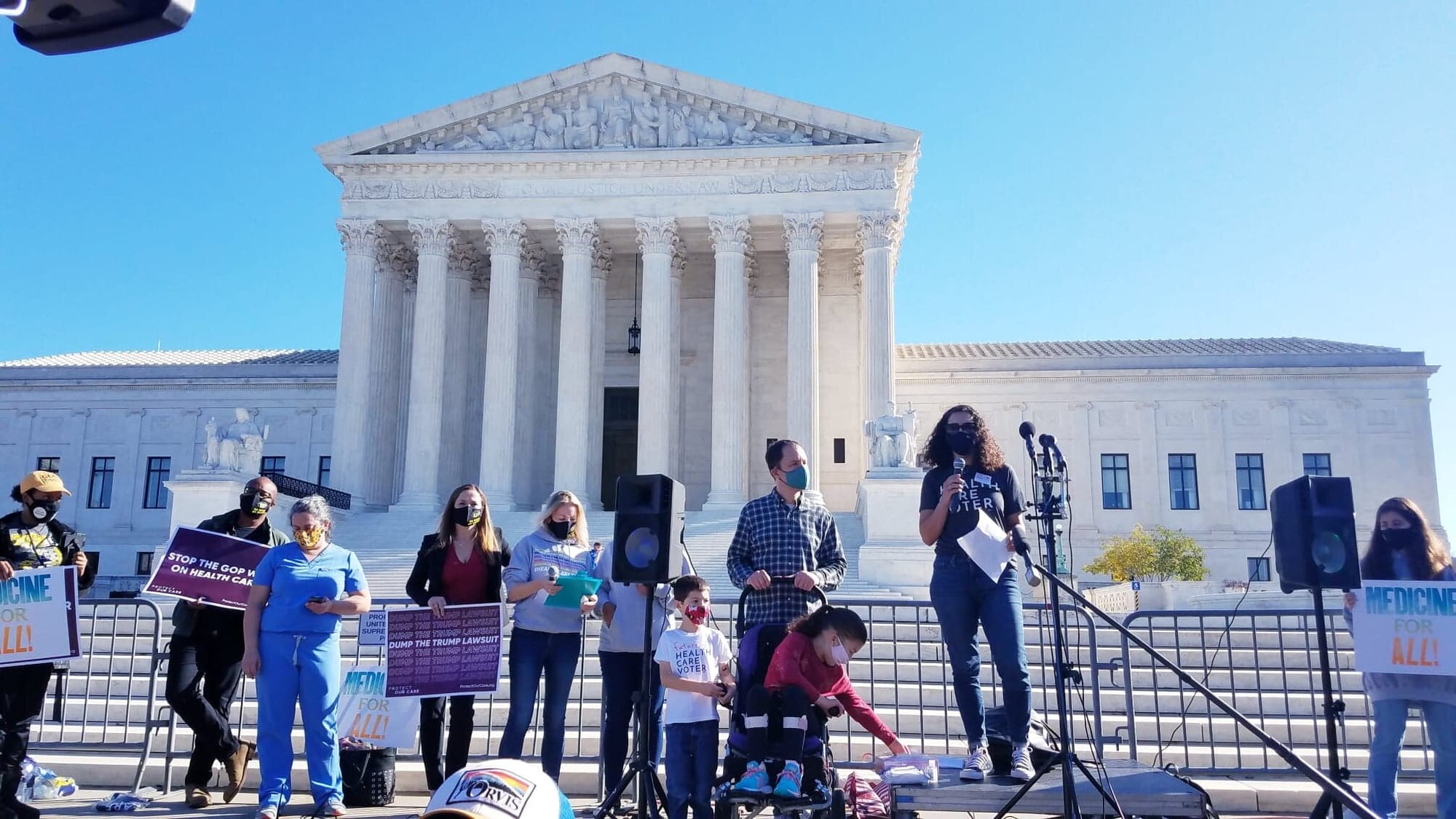 Jamie Davis Smith speaks in front of the Supreme Court. [image description: A woman wearing a face mask stands in front of a microphone. Protestors carrying signs supportive of health care stand in a line to her left & right. In the background i…