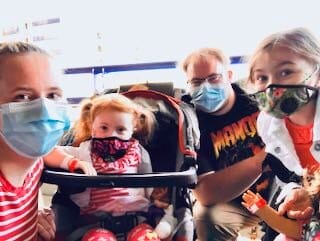 Amanda Upton (l) and her family. [image description: From left to right, a mom, a young daughter, a dad, and another young daughter, all with colorful t-shirts, light hair, and each wearing a face mask, pose outside.]