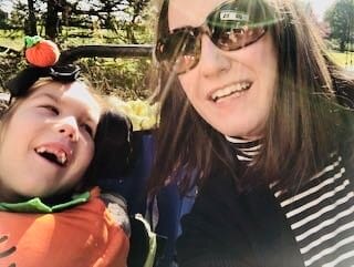 Erin & her daughter Lucia. [image description: A mom with long dark hair, wearing sunglasses, poses with her disabled daughter.