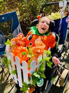 Lucia Raffety-Schneider. [image description: A young disabled girl wears a pumpkin costume for Halloween. Her wheelchair has been turned into a pumpkin patch costume, with a white picket fence, green vines &amp; additional pumpkins.]