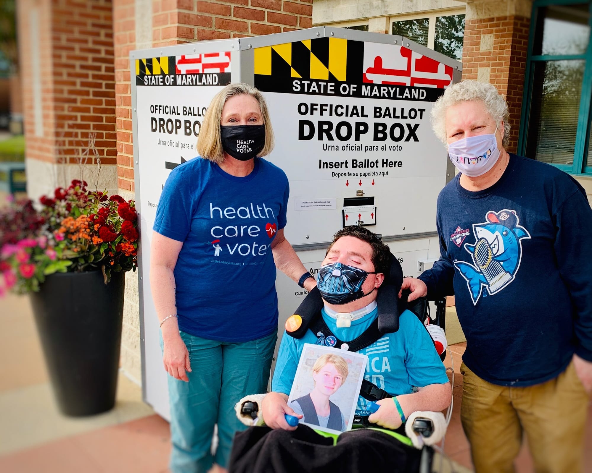 The Stone Family (Jeneva, Rob, Roger) after casting their ballots at a drop box in Maryland. [image description: A woman and two men, one a wheelchair user, pose in front of a State of Maryland Official Drop Box with the colorful logo of the Marylan…