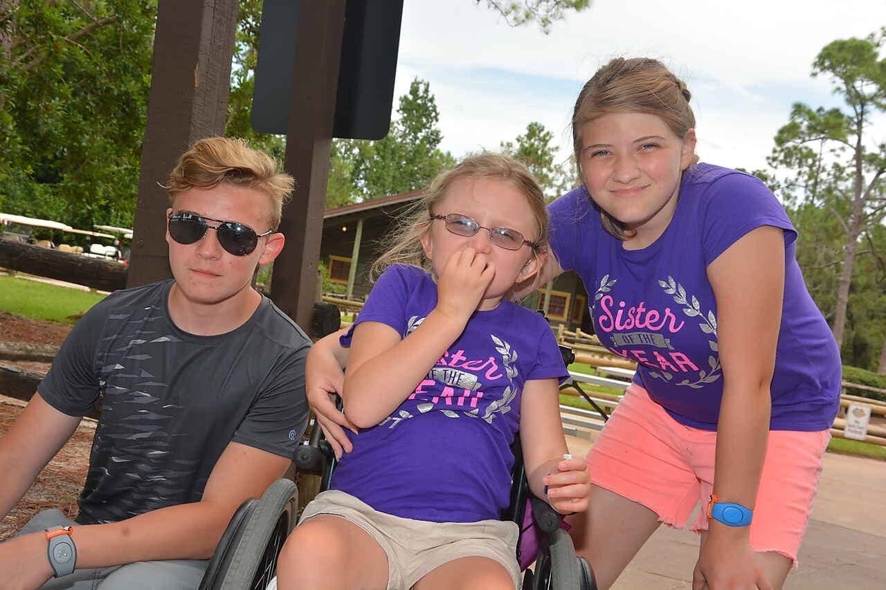 Erin’s three children: Collin, Abby, and Bridget. Collin has short blond hair and wears sunglasses and a gray t-shirt. Abby and Bridget have their blond hair pulled back, and each wears a purple t-shirt with pink lettering that says, “Sister of the …
