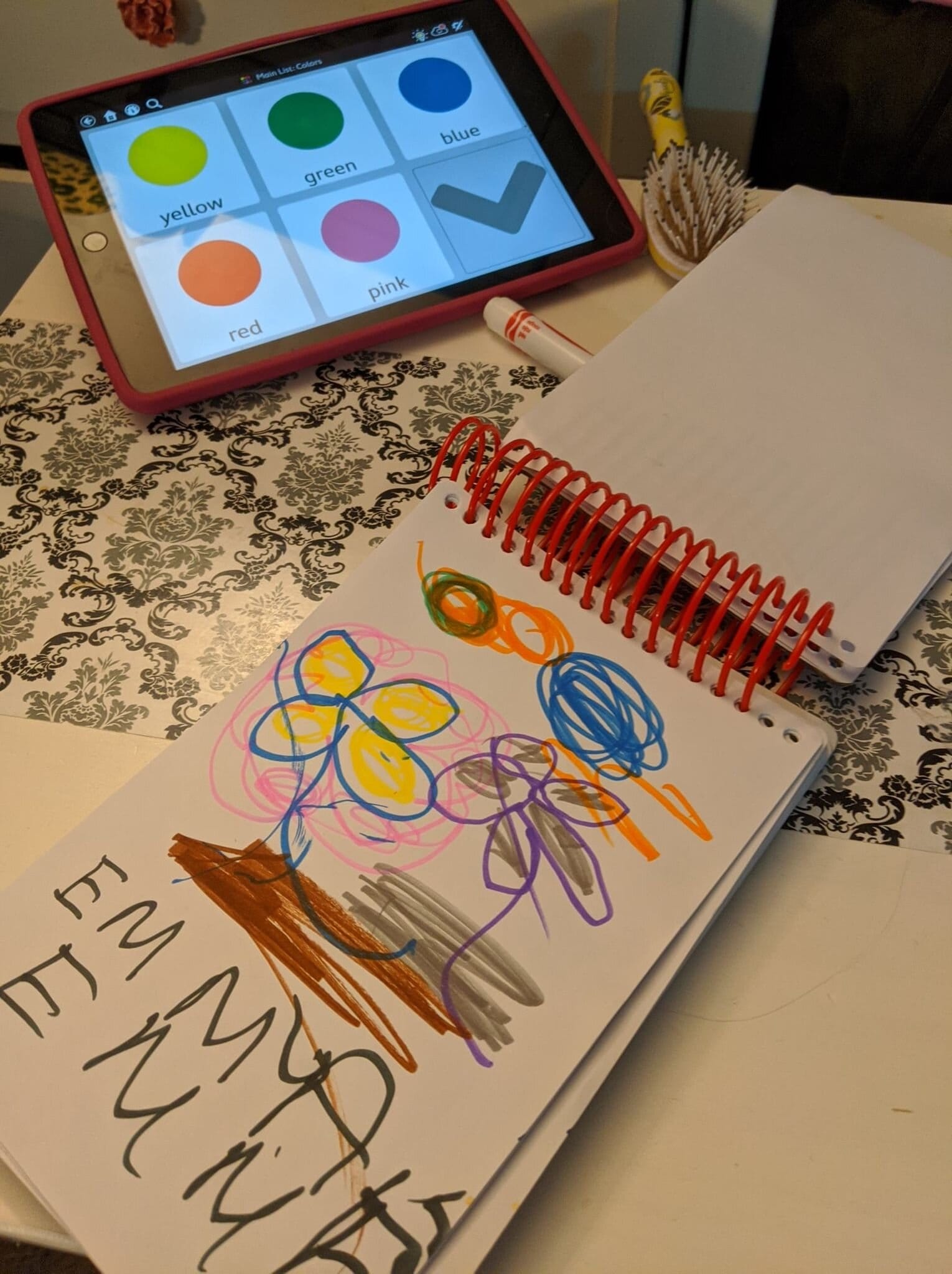 Emma’s AAC device, which resembles an iPad and is in a pink case. The screen displays icons for various colors, and below it is a sketch pad on which Emma has drawn colorful flowers with markers and written her name.