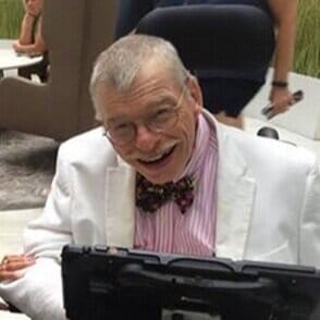 Bob Williams seated in his wheelchair, which has a mount supporting his communication device. Bob is a white man wearing a white suit jacket with a pink striped shirt and a bowtie.