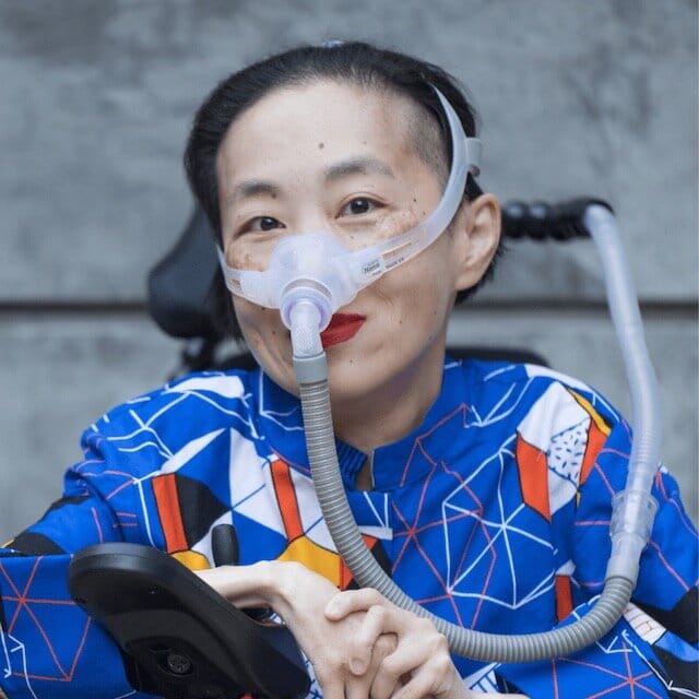 Photo of Alice Wong, an Asian American woman in a power chair. She is wearing a blue shirt with a geometric pattern with orange, black, white, and yellow lines and cubes. She is wearing a mask over her nose attached to a gray tube and bright red lip…
