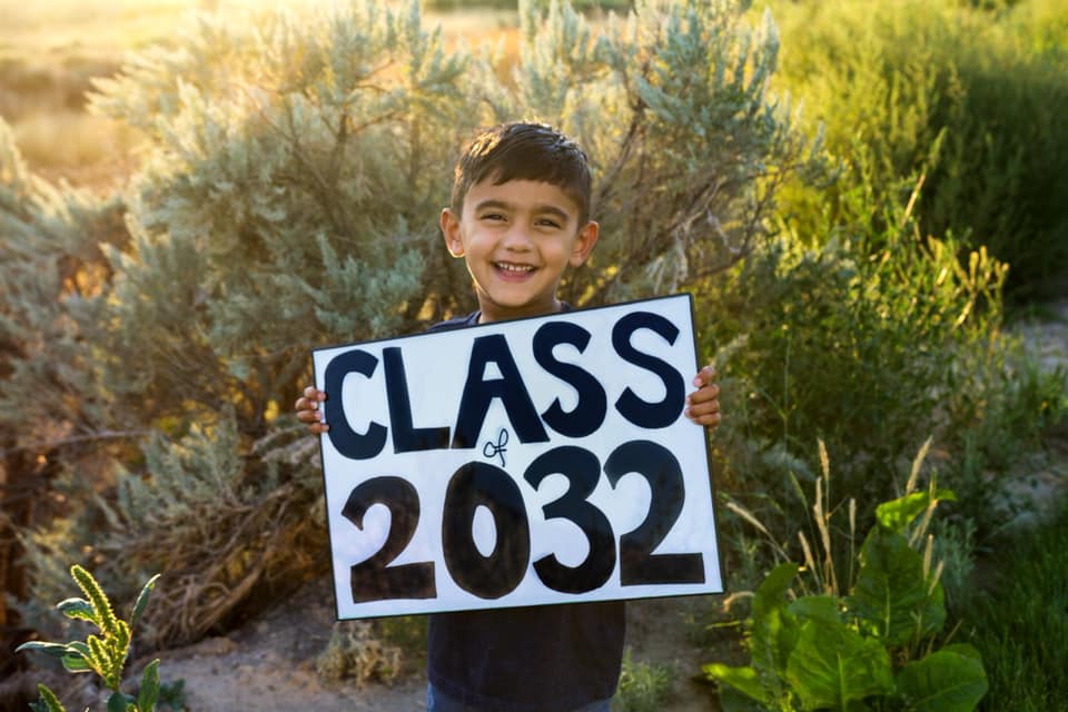 Photo of a smiling little boy standing in front of greenery holding a handmade “Class of 2032” sign.