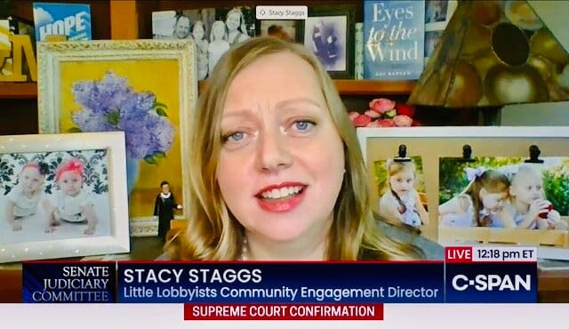 Stacy Staggs speaking before the Senate Judiciary Committee. [Image description: A white woman with blond hair is shown against an array of photos of two young girls, her daughters. A chyron below her states in white print against a blue and red bac…