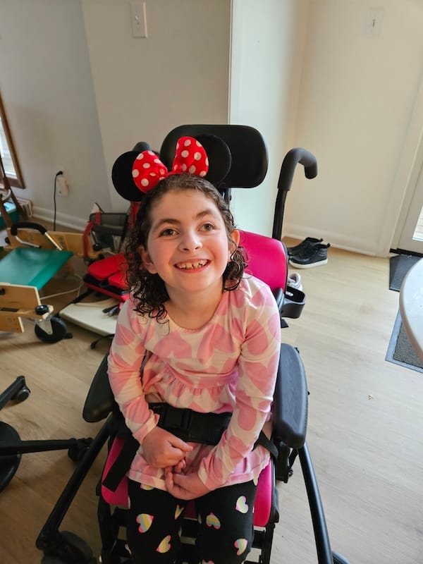 Liviana beaming in Minnie Mouse ears as she sits in her medical stroller.