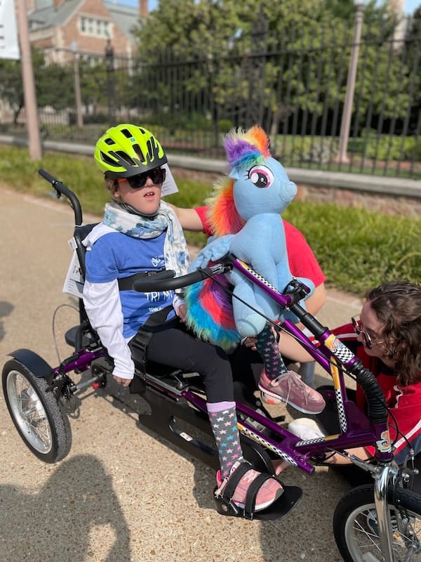 Olive and a stuffed toy riding an adaptive bike.