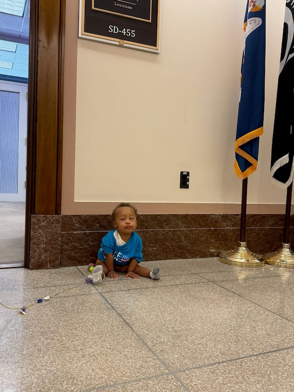 A Black toddler sitting on the floor outside a Congressional office