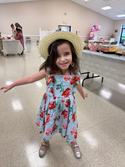Kinsley in a floral dress, stray hat, and silver shoes. She is grinning.