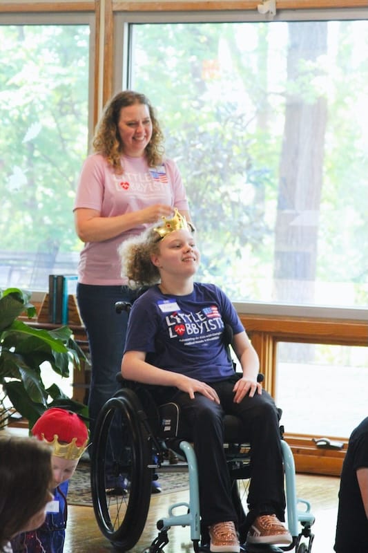 A mother in a Little Lobbyists shirt beaming behind her daughter, who is seated in a wheelchair.