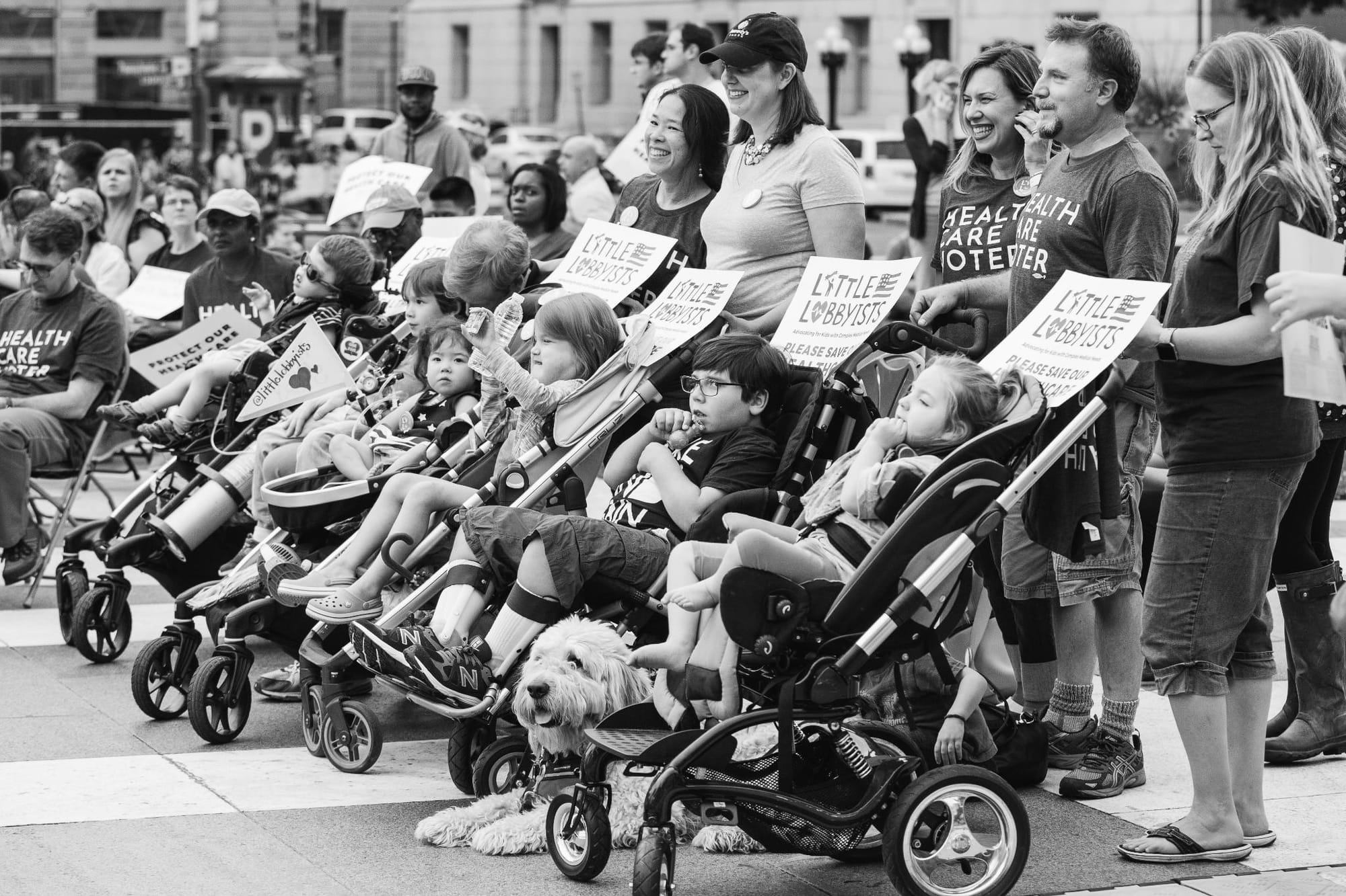 Black-and-white photo of a Little Lobbyists rally on a city street. A row of children sits in strollers and adaptive strollers, holding 'Little Lobbyists' signs that read 'Advocating for Kids with Complex Medical Needs' and 'Please Save Our Healthcare.' Parents stand behind them, some smiling and some wearing 'Healthcare Voter' t-shirts. A shaggy dog rests on the ground in front of the strollers. A larger crowd of demonstrators and city buildings fill the background.