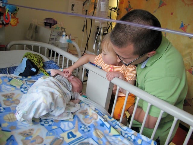 James in his crib in a hospital setting, with his father and sibling leaning in to hold his hand.