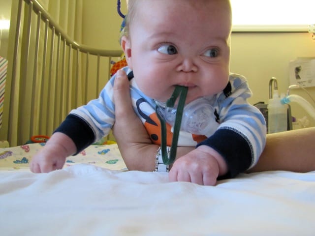 James on his tummy in his crib, looking slightly worried. His tracheostomy is visible.