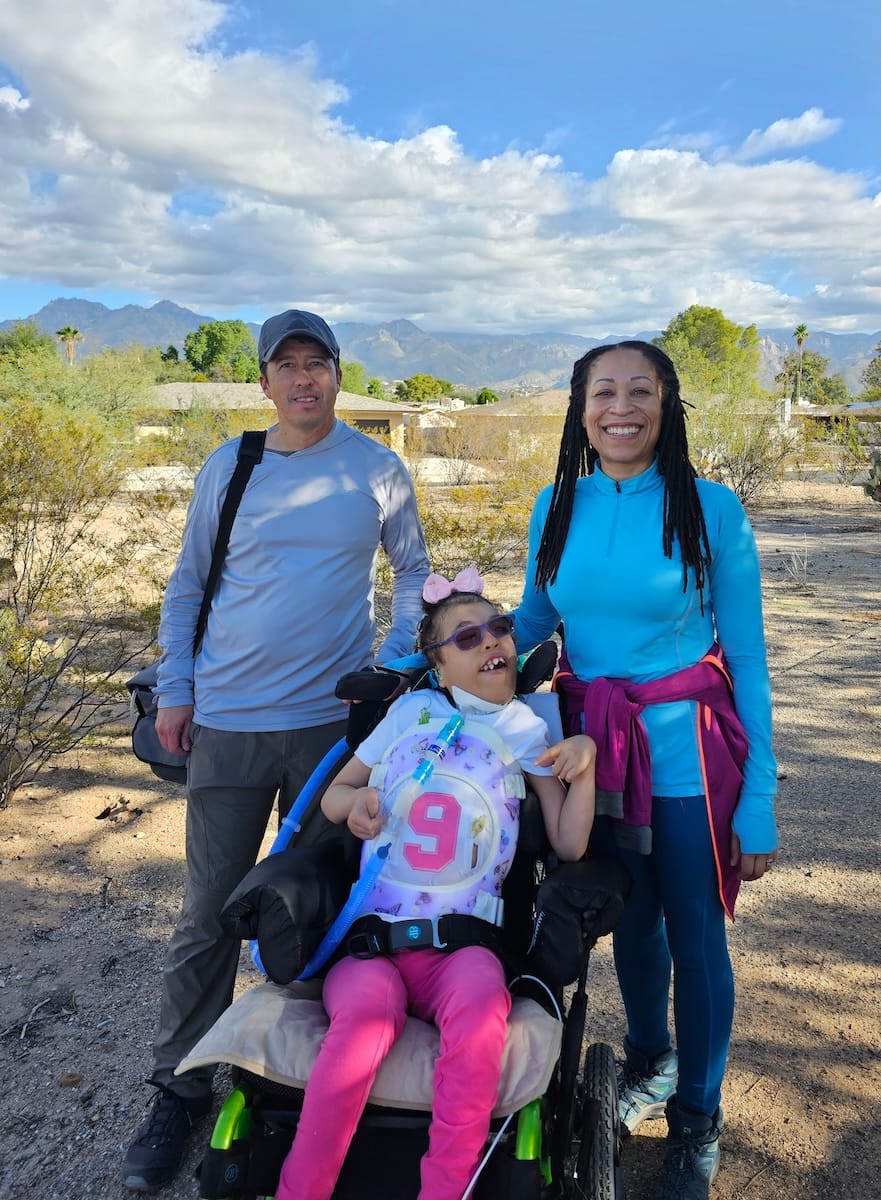 Dianne, a Black woman with long hair; Matt, a white man; and Madison smiling for a family photo outside on a beautiful day.