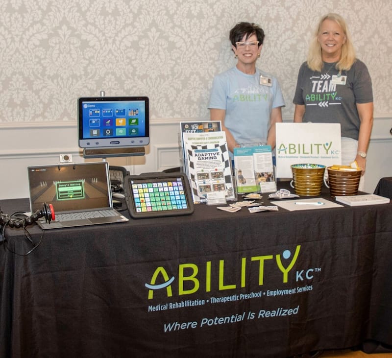 Two women smiling as they demonstrate various AAC devices.