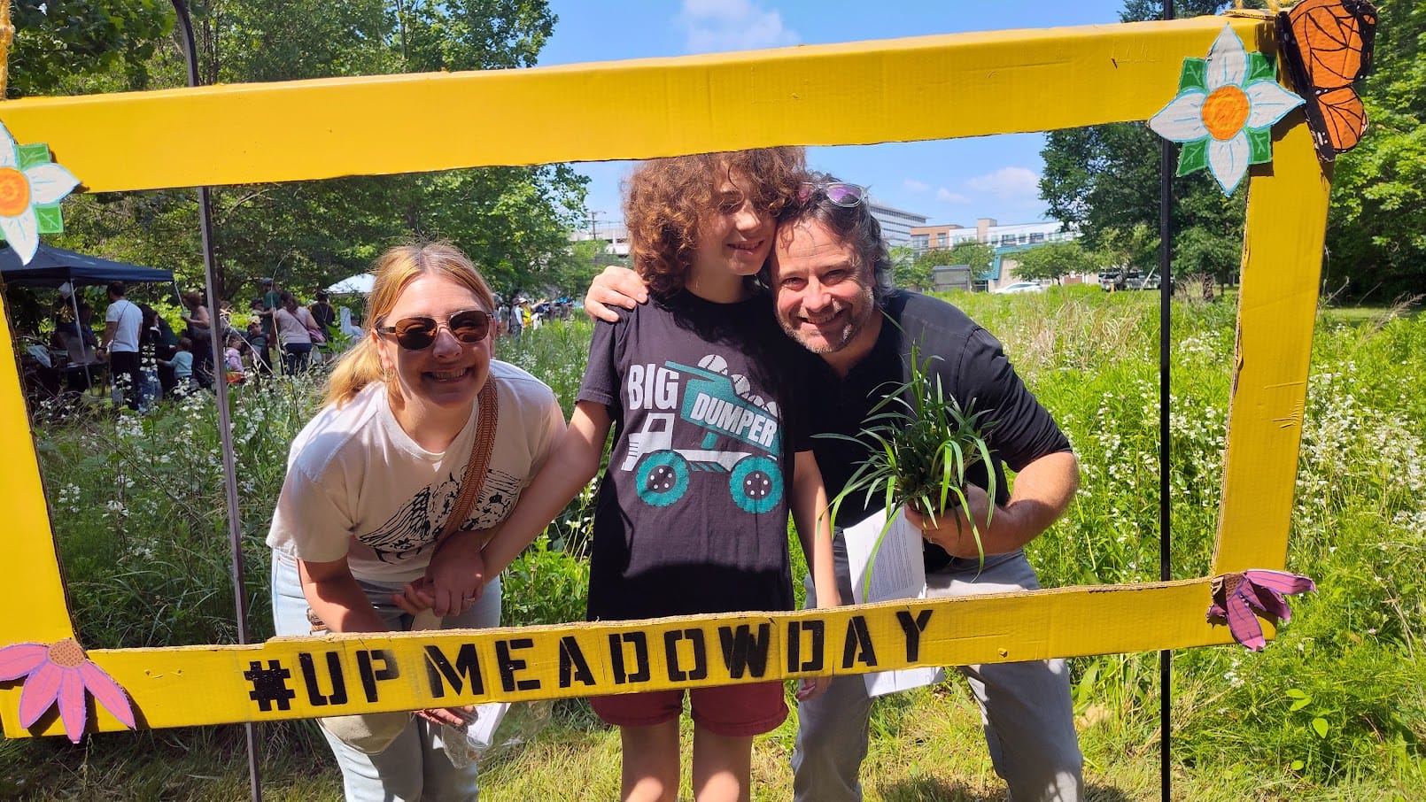 Lazaro and his family smiling outside in a beautiful meadow from within a cardboard frame labeled #UpMeadowDay