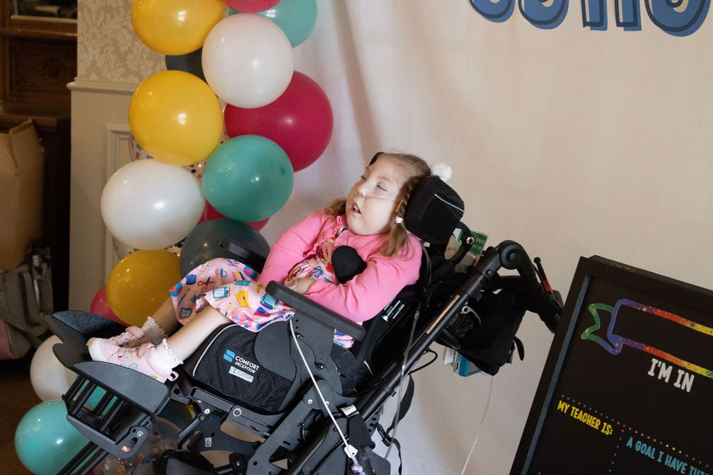 A white child sitting in her wheelchair next to a cluster of balloons.
