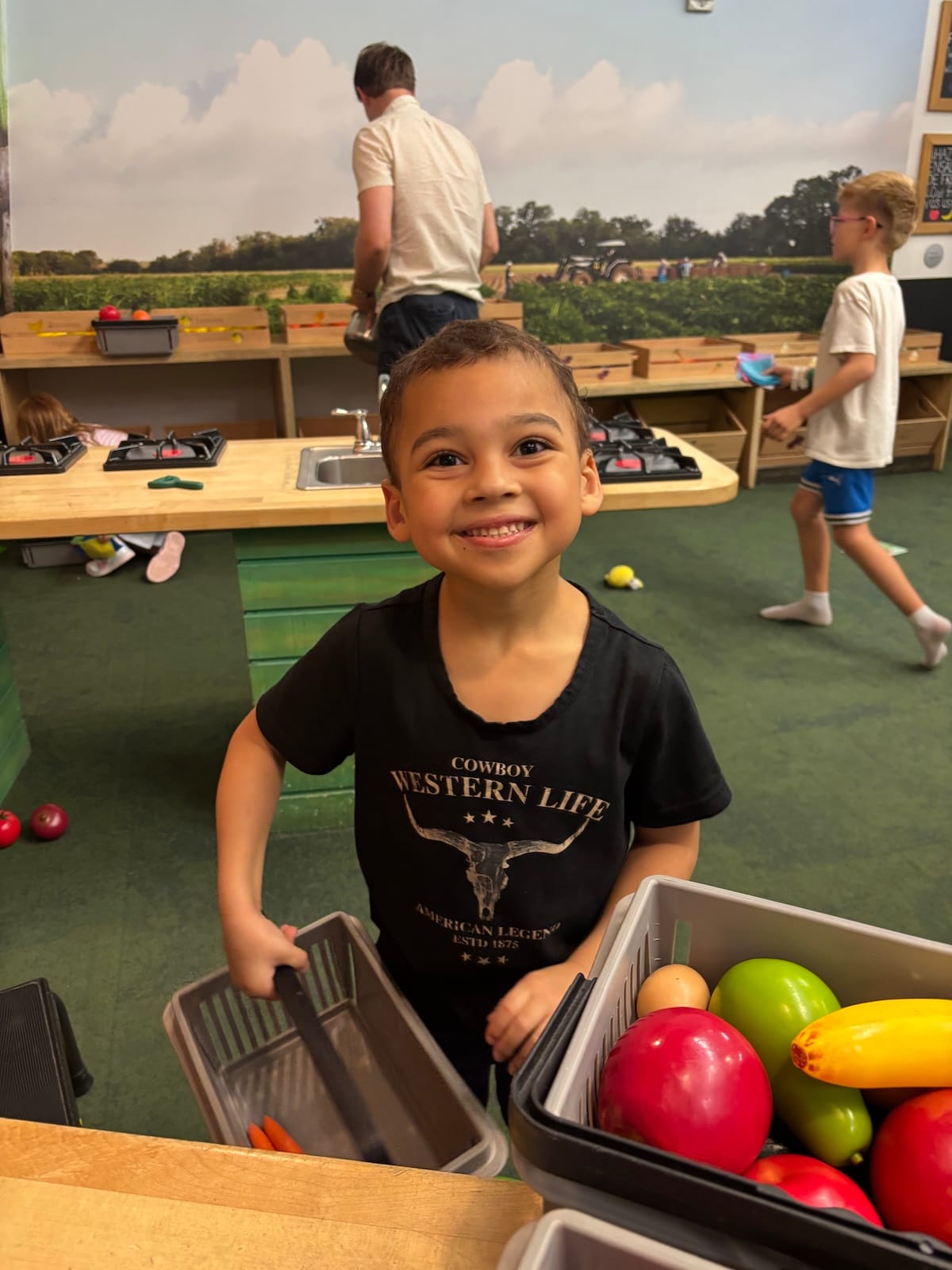 JW, a biracial child with a huge smile, plays with plastic fruit in a classroom.