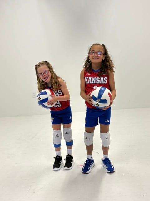 Two girls, both in volleyball uniforms with balls, smiling for the camera.