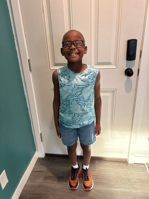 A Black child with a shaved head and big glasses, wearing shorts and a tank top, with a huge smile.