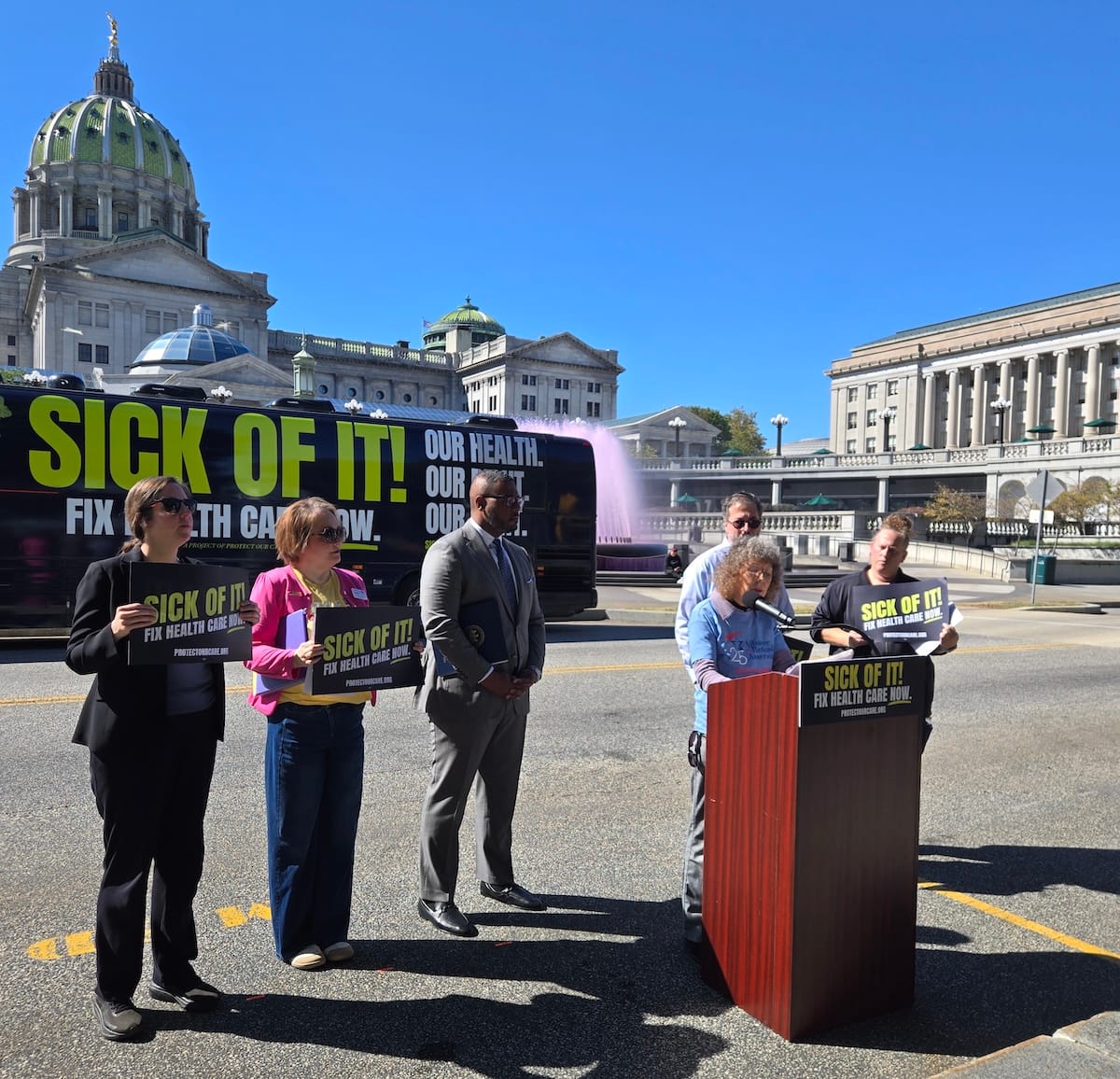 A white woman with a cloud of curly hair speaking, surrounded by advocates with Sick of It! Fix Health Care Now! signs.