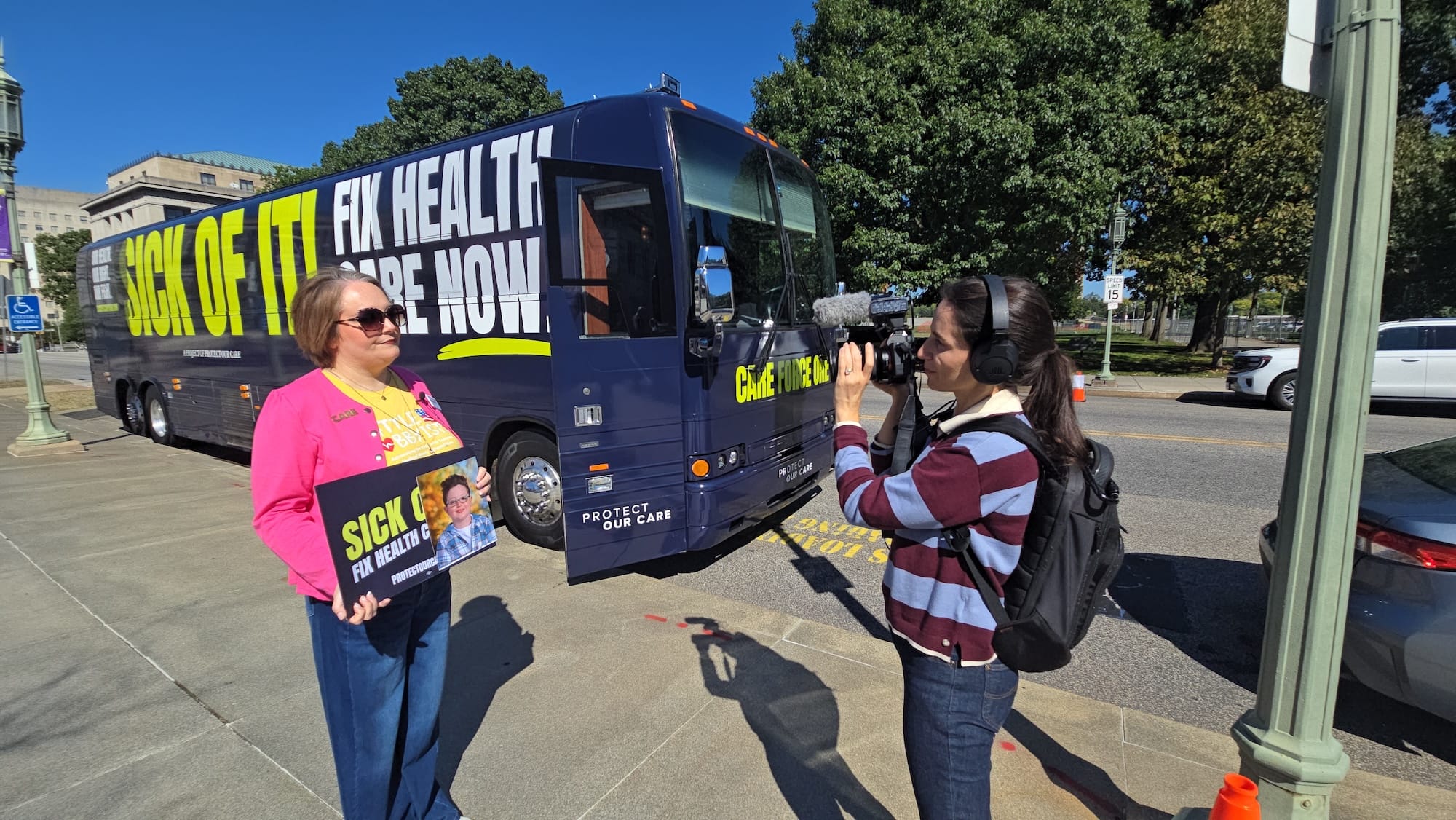 Victoria Schiano, a white woman with a bob, holds up a photo of her son Cole in front of the Care Force One bus while being interviewed.